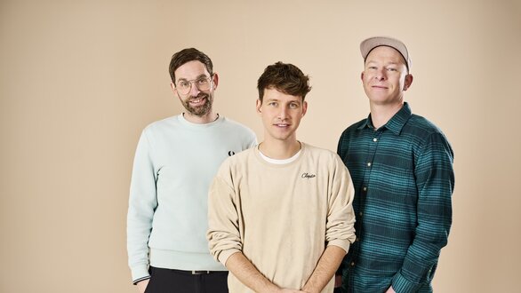 Drei junge Männer stehen lächelnd vor einer neutralen Wand.