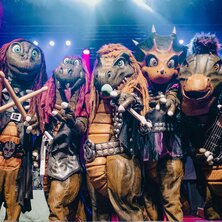 Fünf scurrile Figuren in Dinosaurierkostümen posieren auf einer Bühne.