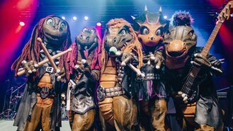 Fünf scurrile Figuren in Dinosaurierkostümen posieren auf einer Bühne.