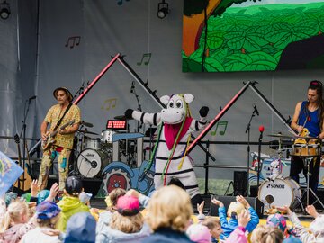 Eine kindgerechte Band mit einem Maskottchen tritt auf einer Bühne auf.