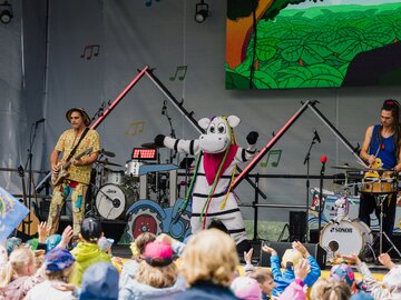 Eine kindgerechte Band mit einem Maskottchen tritt auf einer Bühne auf.