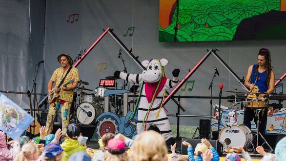 Eine kindgerechte Band mit einem Maskottchen tritt auf einer Bühne auf.