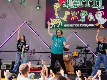 Drei Performer tanzen auf einer Bühne vor Kindern mit Animation.