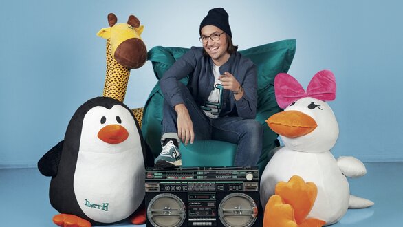 Ein Moderator sitzt zwischen einem Pinguin und einer Giraffe auf einem Sofa.