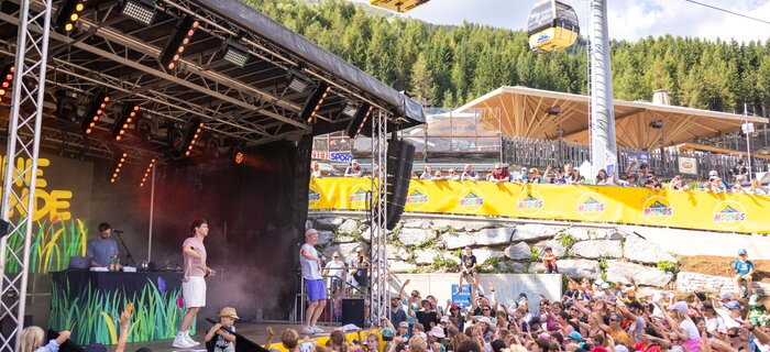 Bühne mit DJ und tanzenden Menschen unter einer Gondel.