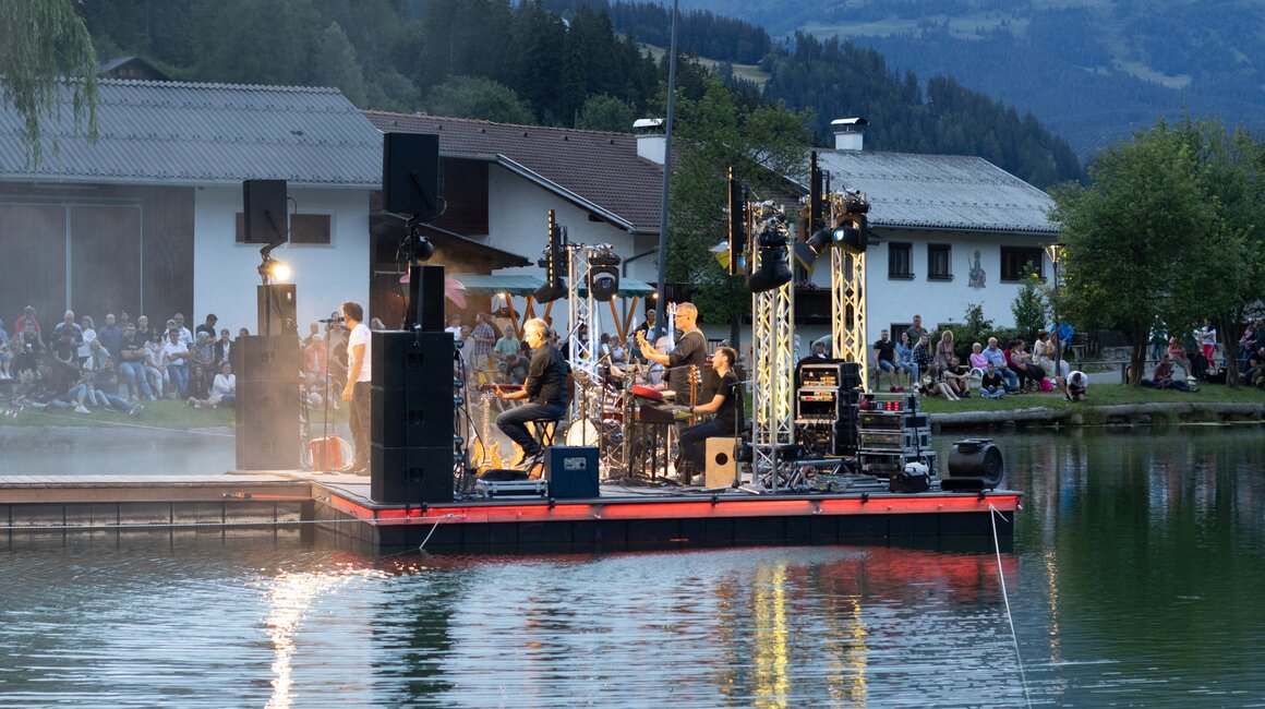 Eine Band spielt auf einer schwimmenden Bühne, umgeben von Zuschauern am Ufer.
