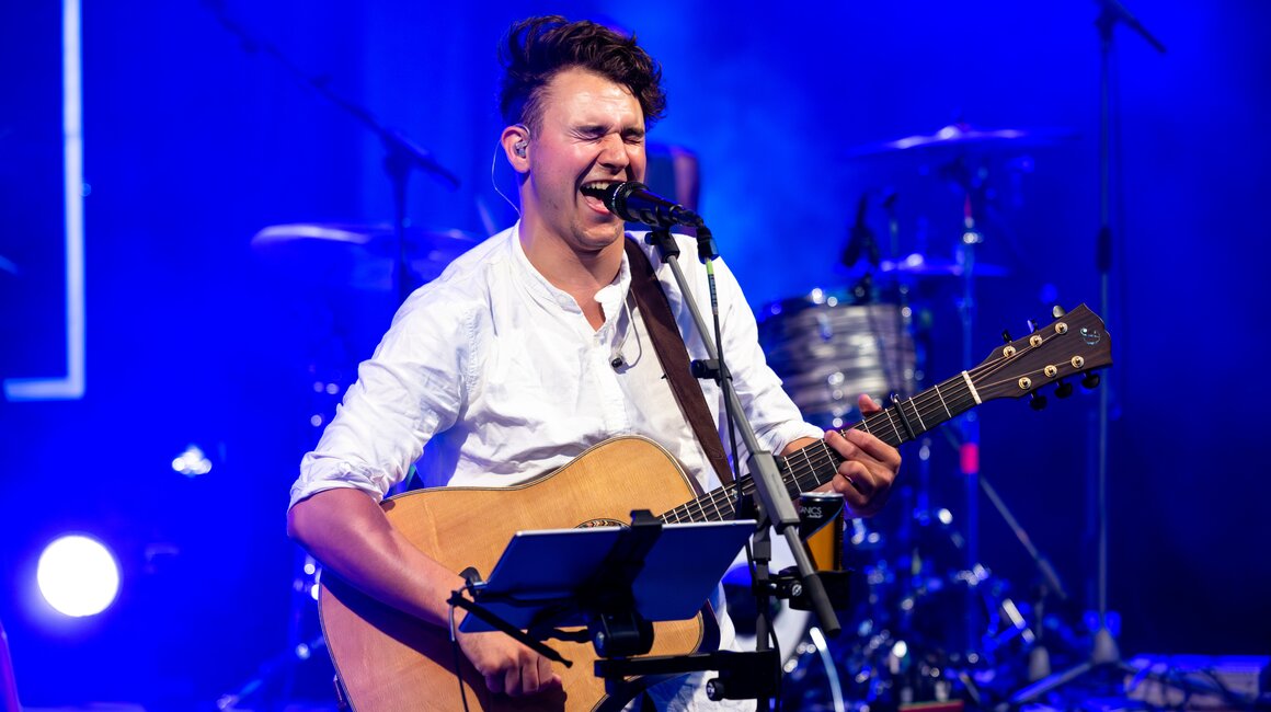 Ein Sänger spielt Gitarre und singt leidenschaftlich auf einer beleuchteten Bühne.