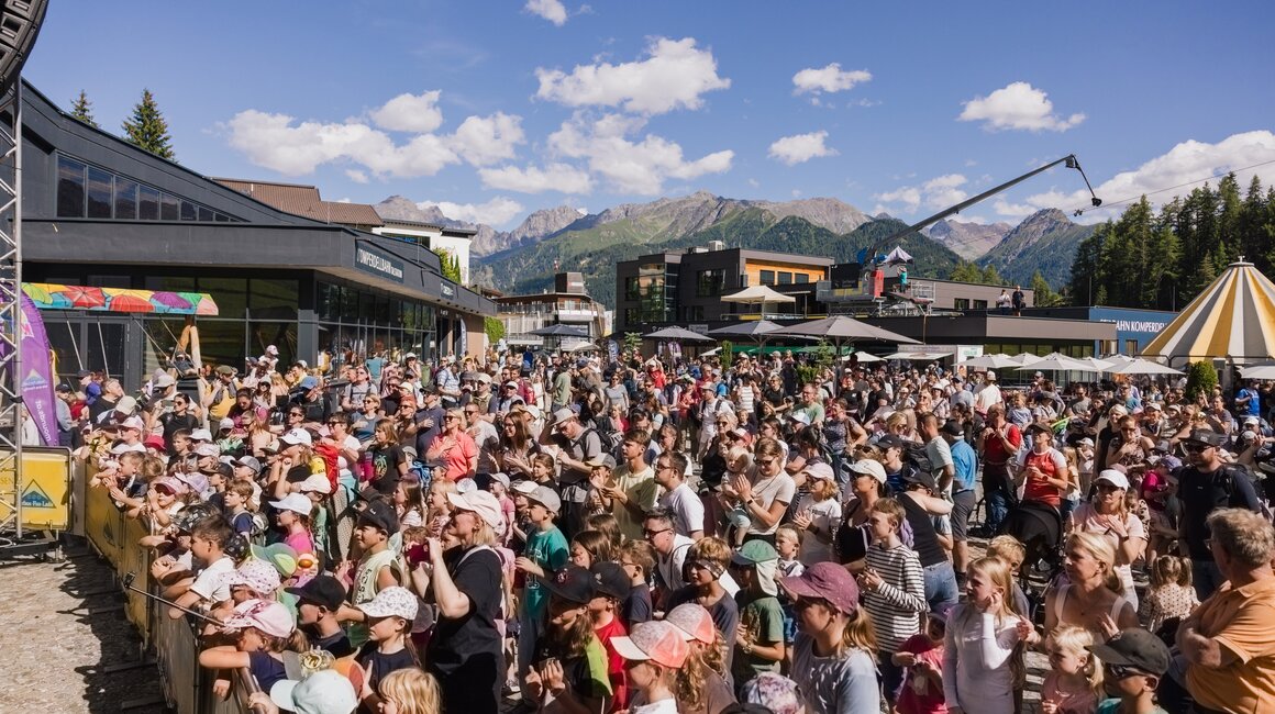 Eine große Menschenmenge bei einem Fest im Freien mit Bergen im Hintergrund.