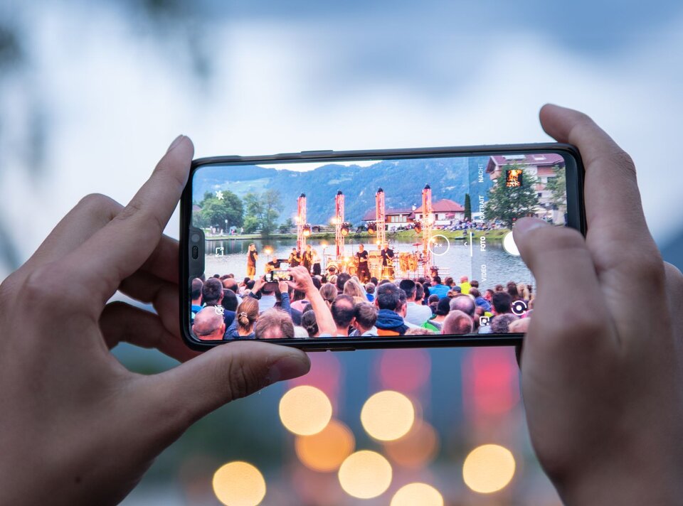 Eine Person filmt eine Show mit ihrem Handy am See, umgeben von Publikum.